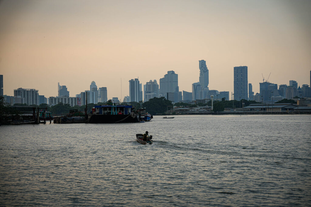 Ein kleines Boot mit zwei Personen fährt in der Abenddämmerung über einen Fluss in Thailand. Im Hintergrund zeichnen sich die Silhouette einer Stadt und hohe Gebäude in einem dunstigen Himmel ab.