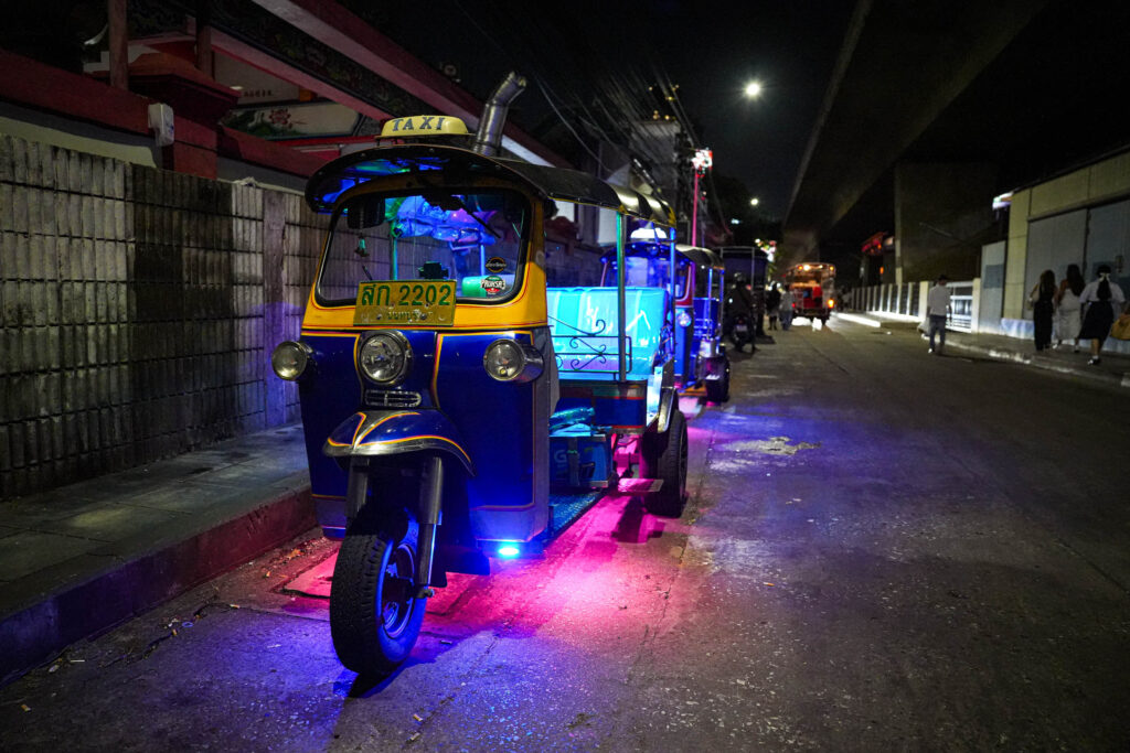 Ein hell erleuchtetes Tuk-Tuk-Taxi mit Neonröhren parkt nachts auf einer Stadtstraße in Thailand, dahinter reihen sich weitere Tuk-Tuks. Die farbenfrohe Szene fängt das pulsierende Nachtleben ein, das viele zum Auswandern nach Thailand anzieht.