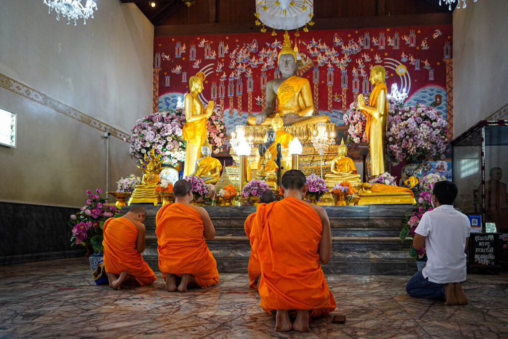 Fünf Mönche in orangefarbenen Gewändern und ein Laie knien und beten vor einer goldenen Buddha-Statue in einem reich geschmückten thailändischen Tempel, der mit Blumen und roten Ornamenten verziert ist - und das spirituelle Herz derjenigen einfängt, die nach Thailand auswandern wollen.