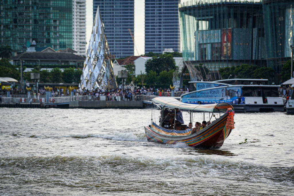 Ein farbenfrohes Longtail-Boot mit Passagieren fährt auf einem Fluss in Thailand, im Hintergrund sind ein geschmückter silberner Weihnachtsbaum und moderne Glasgebäude am belebten Flussufer zu sehen.