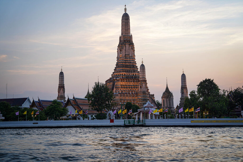 Der Wat Arun Tempel steht majestätisch am Chao Phraya Fluss in Bangkok, wenn die Sonne untergeht. Seine verschnörkelten Türme leuchten gegen den Himmel - eine atemberaubende Szene, die viele dazu inspiriert, ein neues Abenteuer in Thailand zu wagen.