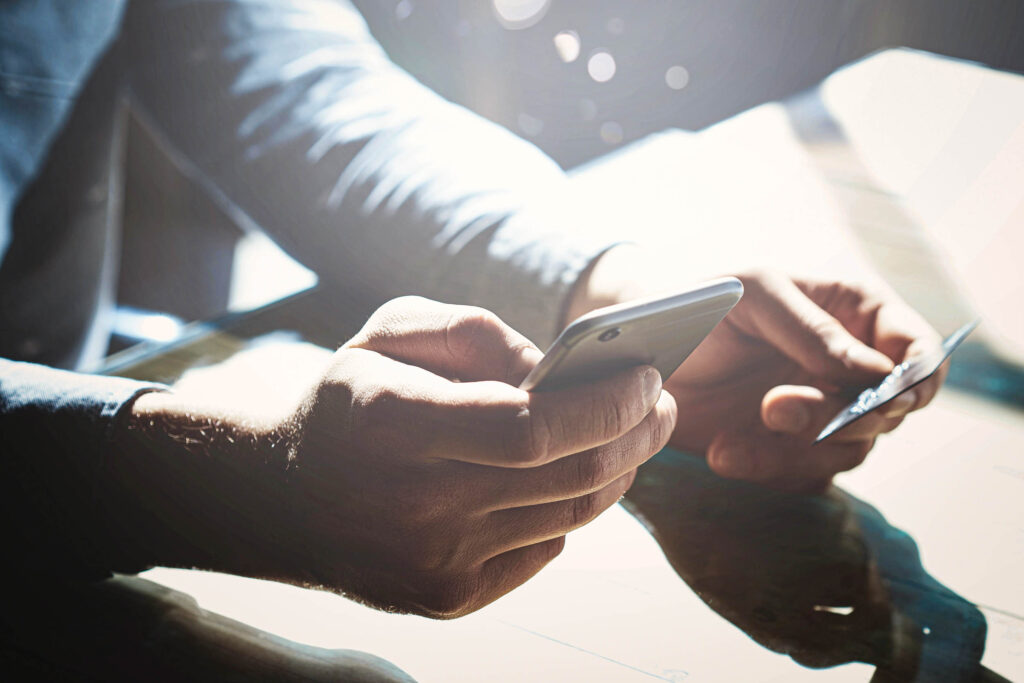 Eine Person, die an einem Tisch sitzt, hält in der einen Hand ein Smartphone und in der anderen eine Kreditkarte, während das Sonnenlicht auf die Oberfläche fällt.