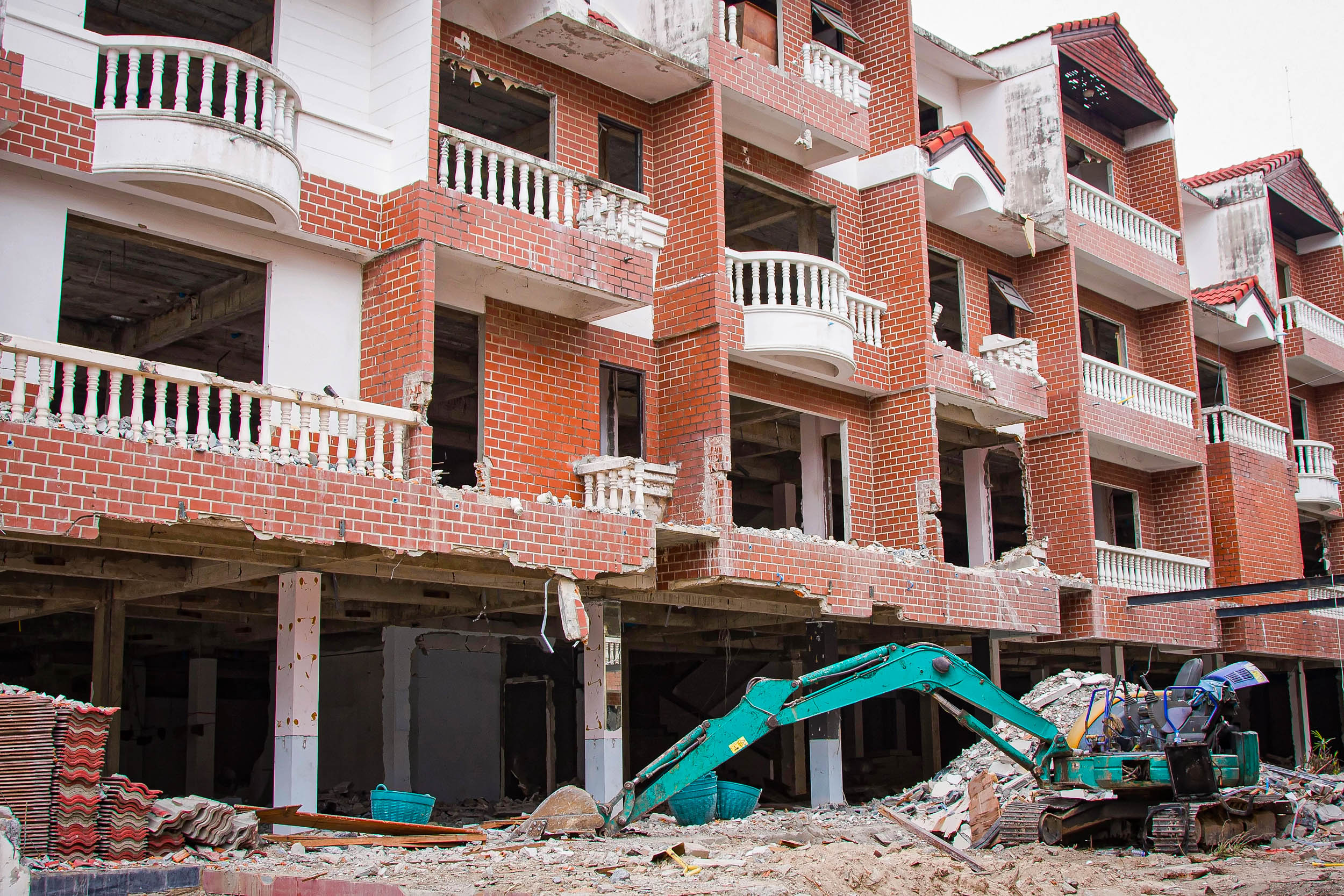 Ein teilweise abgerissenes rotes Backsteingebäude mit freigelegten Böden und Wänden. Davor steht ein grüner Bagger, umgeben von Schutt und Baumaterial. Einige Balkone und Strukturelemente sind noch intakt.