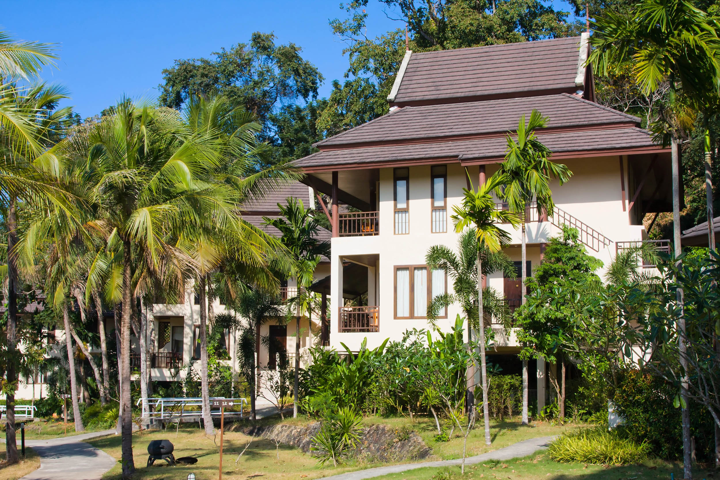 Zwei cremefarbene, moderne tropische Villen mit Holzdächern und Balkonen sind von Palmen und üppigem Grün unter einem klaren blauen Himmel umgeben.