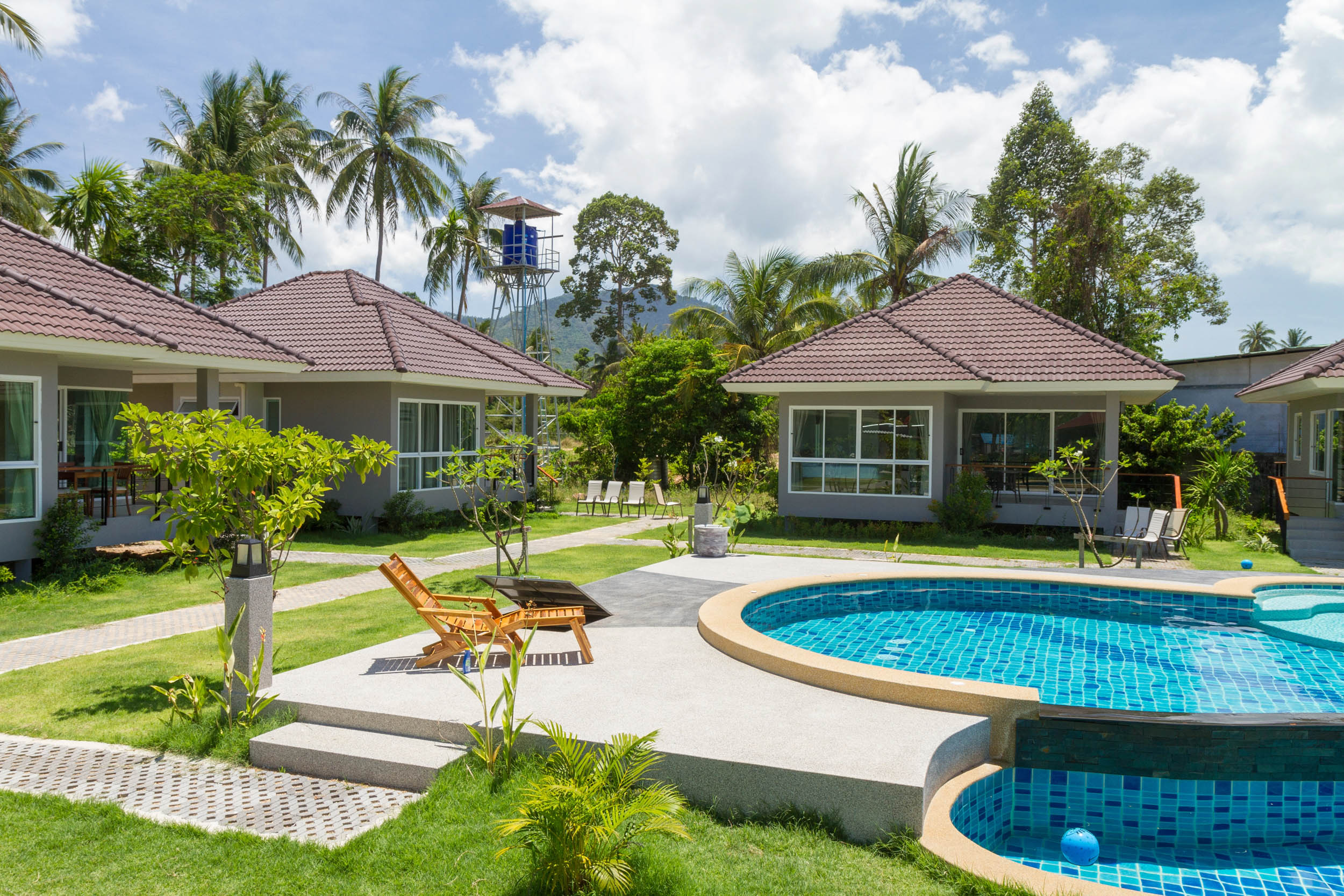 Eine tropische Resort-Szene mit mehreren Bungalows, einem runden Swimmingpool, Sonnenliegen am Pool, grünen Rasenflächen und Palmen unter einem strahlend blauen Himmel mit vereinzelten Wolken.