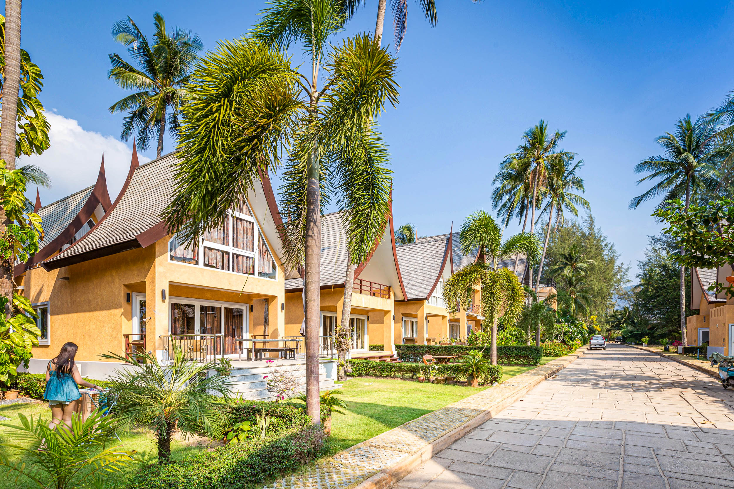 Eine Reihe tropischer Villen mit spitzen Dächern und gelben Mauern, gesäumt von Palmen an einem sonnigen Tag, mit einem Mädchen in einem blauen Kleid, das auf dem Rasen neben dem Gehweg spazieren geht.