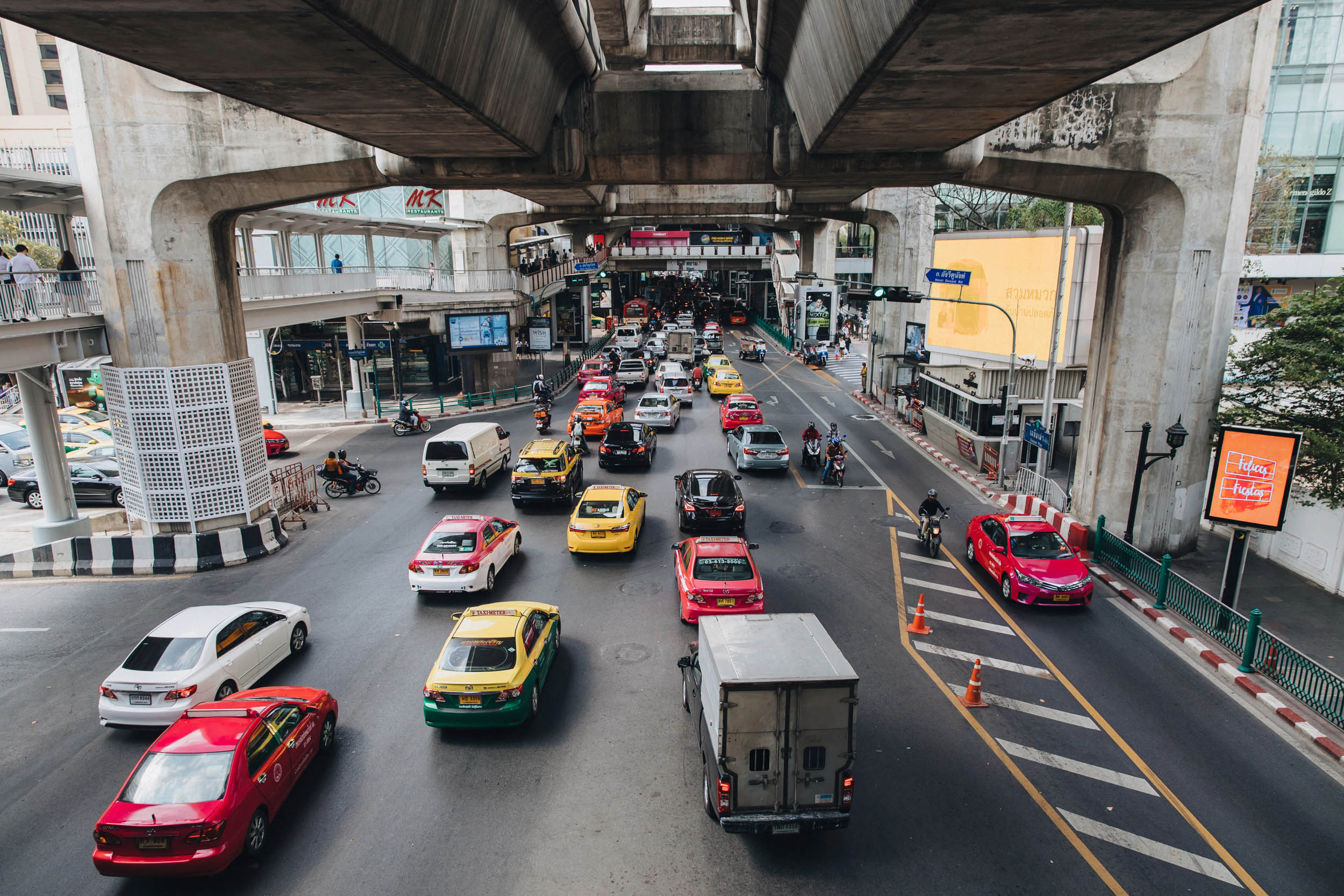 Eine belebte Stadtstraße mit vielen Autos, Taxis und einem Lastwagen, der unter einer Hochbahnstrecke hindurchfährt, umgeben von hohen Gebäuden, Verkehrsschildern und Fußgängern auf den Bürgersteigen.
