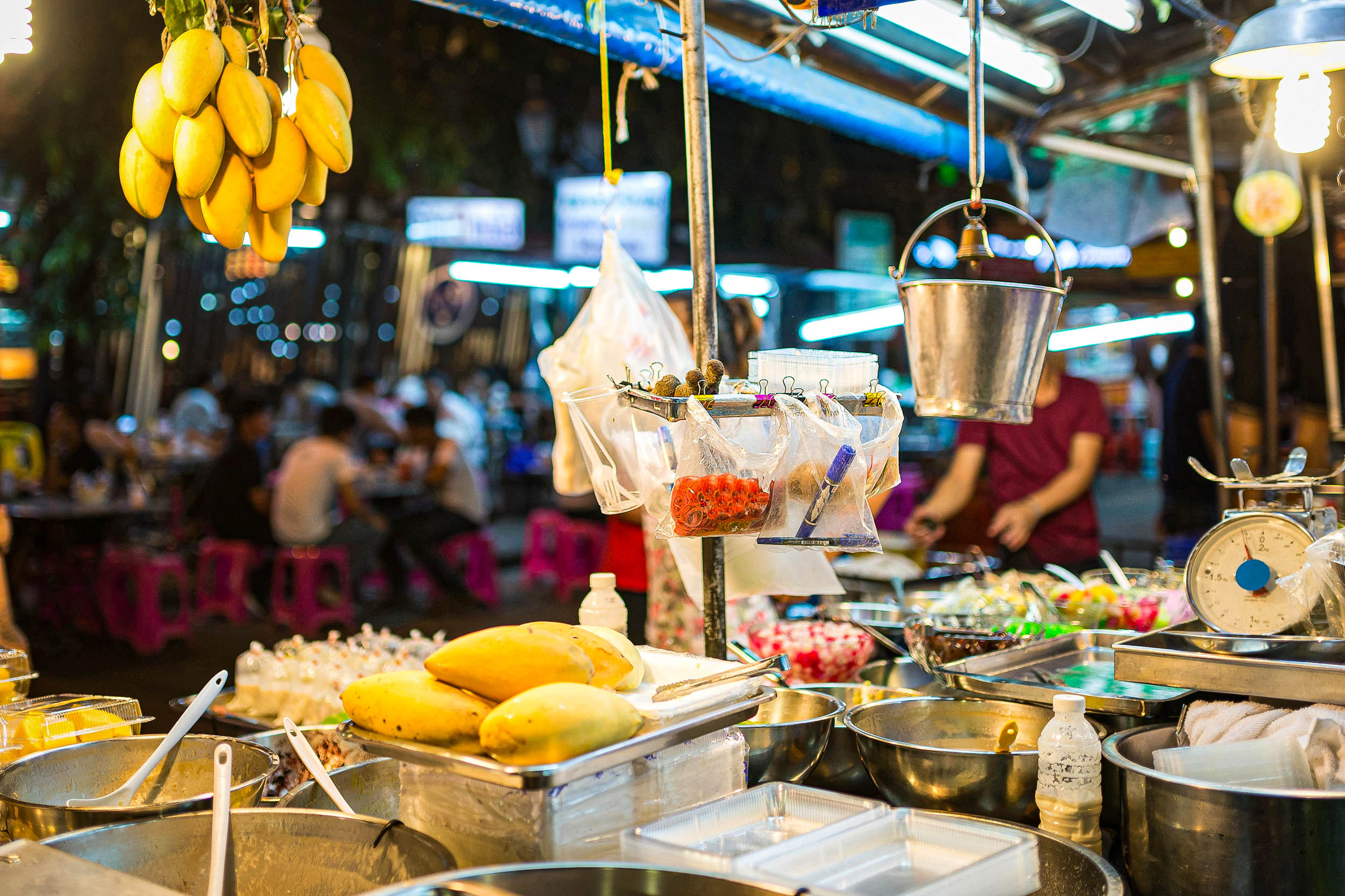 Ein lebhafter Streetfood-Stand bietet frische Mangos, in Plastik verpackte Snacks, Metallschüsseln, eine Waage und einen hängenden Eimer an, während im Hintergrund Menschen an rosa Tischen unter hellen Lichtern essen.