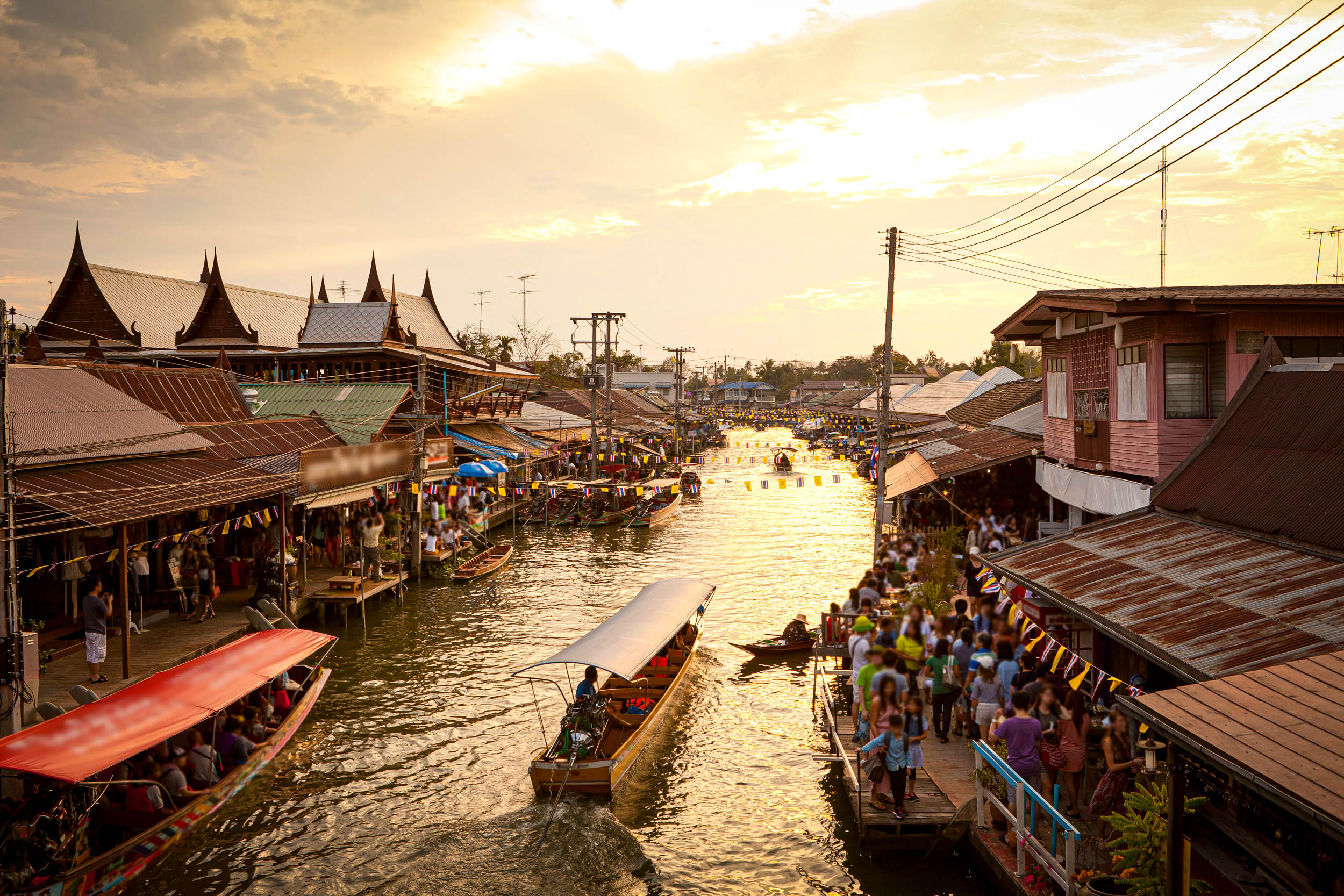 Ein belebter schwimmender Markt bei Sonnenuntergang, mit Holzbooten in einem engen Kanal und Menschen, die sich an beiden Ufern drängen. Traditionelle Häuser säumen die Wasserstraße, und bunte Fahnen hängen über der lebhaften Szene.