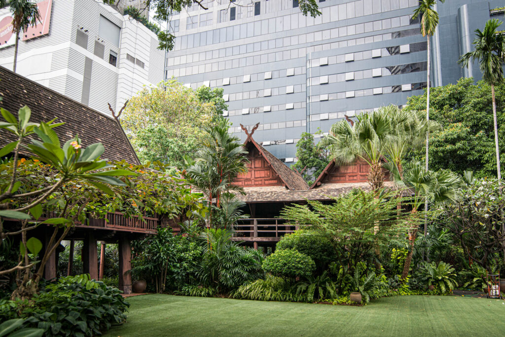 Ein traditionelles thailändisches Holzhaus, umgeben von üppigen tropischen Pflanzen, steht vor modernen Hochhäusern aus Glas und Beton in einer städtischen Umgebung.