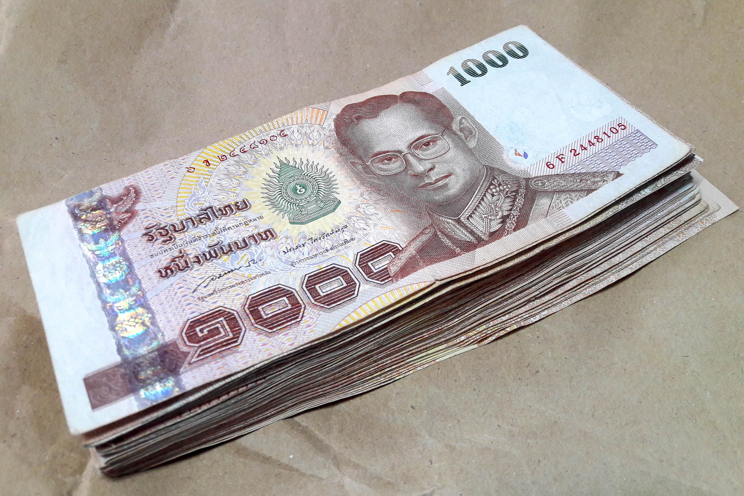 Ein Stapel von 1.000 Thai-Baht-Banknoten auf einer braunen Oberfläche. Die Scheine zeigen das Bild eines Mannes mit Brille und traditioneller Kleidung.