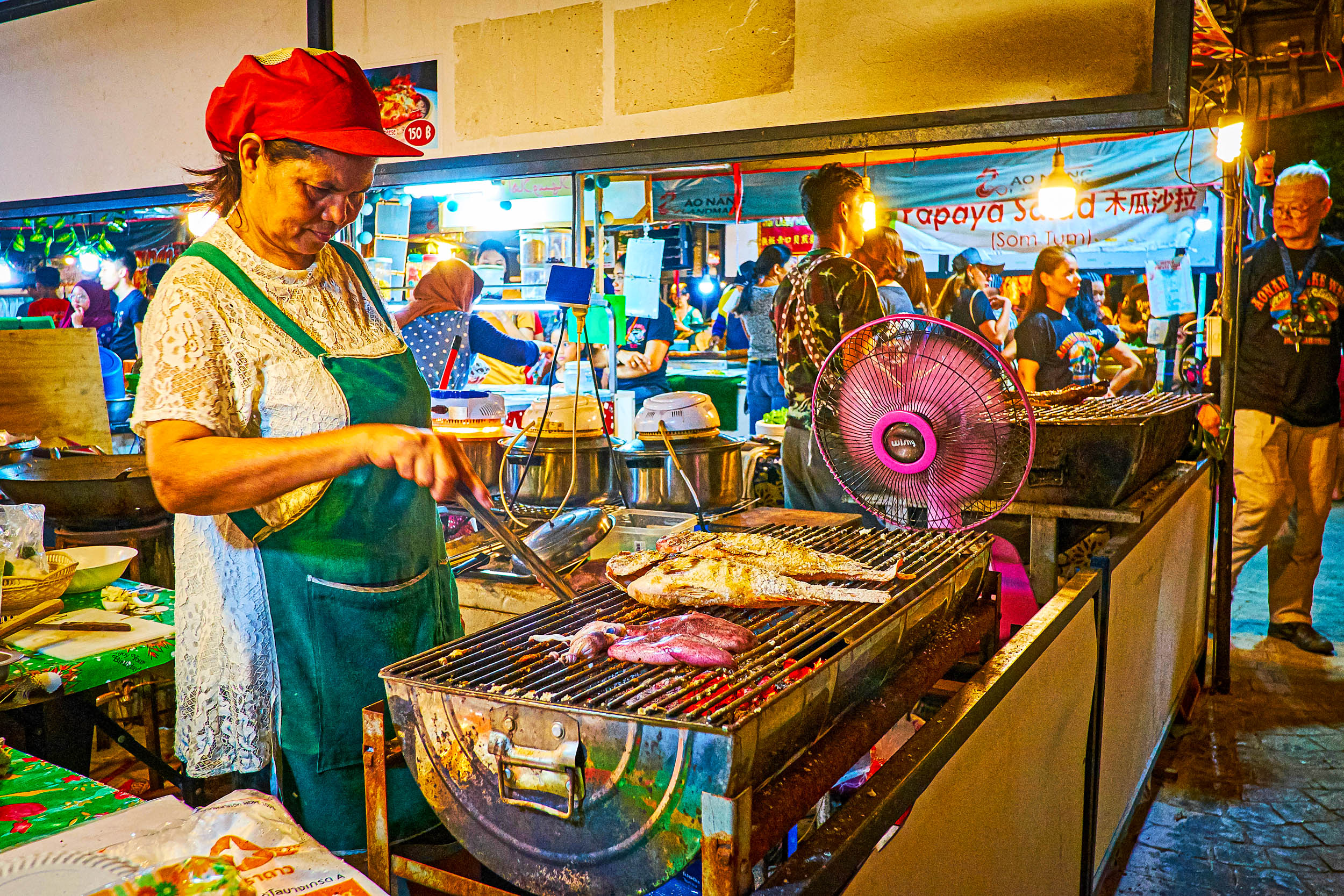 Eine Frau mit rotem Hut und grüner Schürze grillt Fisch an einem belebten Marktstand in einer Halle. Im lebhaften Hintergrund sind bunte Lichter, andere Verkäufer und Kunden zu sehen.