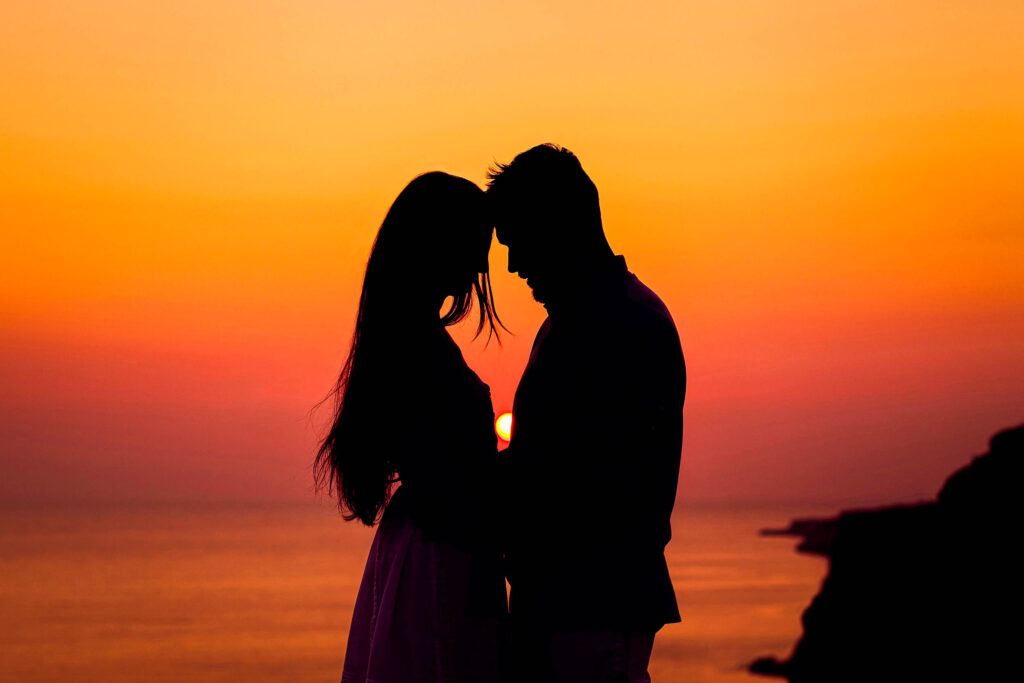 Silhouette eines Paares, das während des Sonnenuntergangs in Thailand eng beieinander am Meer steht, mit einem orange-gelben Himmel im Hintergrund. Die Sonne geht in der Nähe des Horizonts unter und wirft ein warmes Licht auf das Wasser - perfekt für alle, die vom Auswandern träumen.