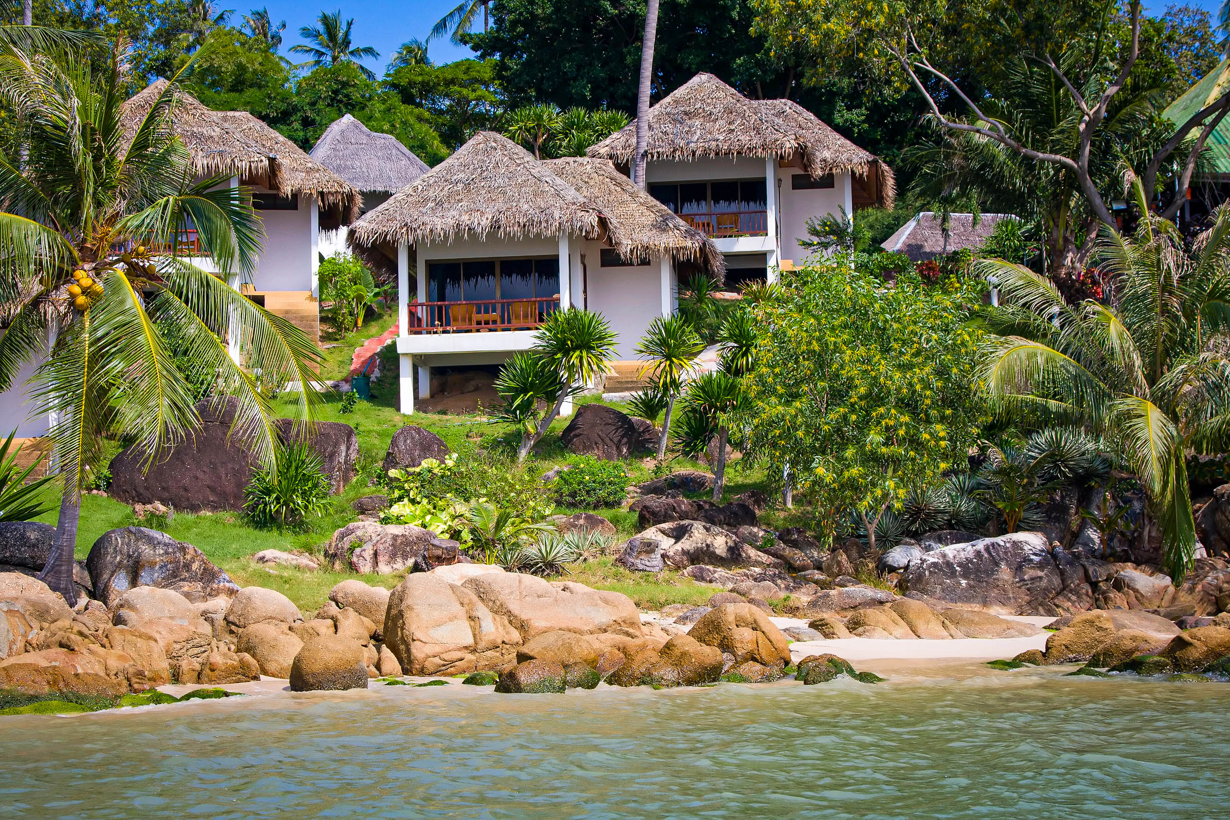 Tropische Bungalows mit Strohdächern am Strand, umgeben von üppigem Grün, Palmen, Felsen und Sandstrand, vom Wasser aus gesehen.