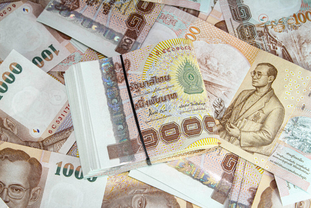 Ein Bündel thailändischer Baht-Banknoten, die mit einem schwarzen Band gesichert sind, ruht auf verstreuten thailändischen 1000-Baht-Noten, die alle das Porträt von König Bhumibol Adulyadej zeigen.