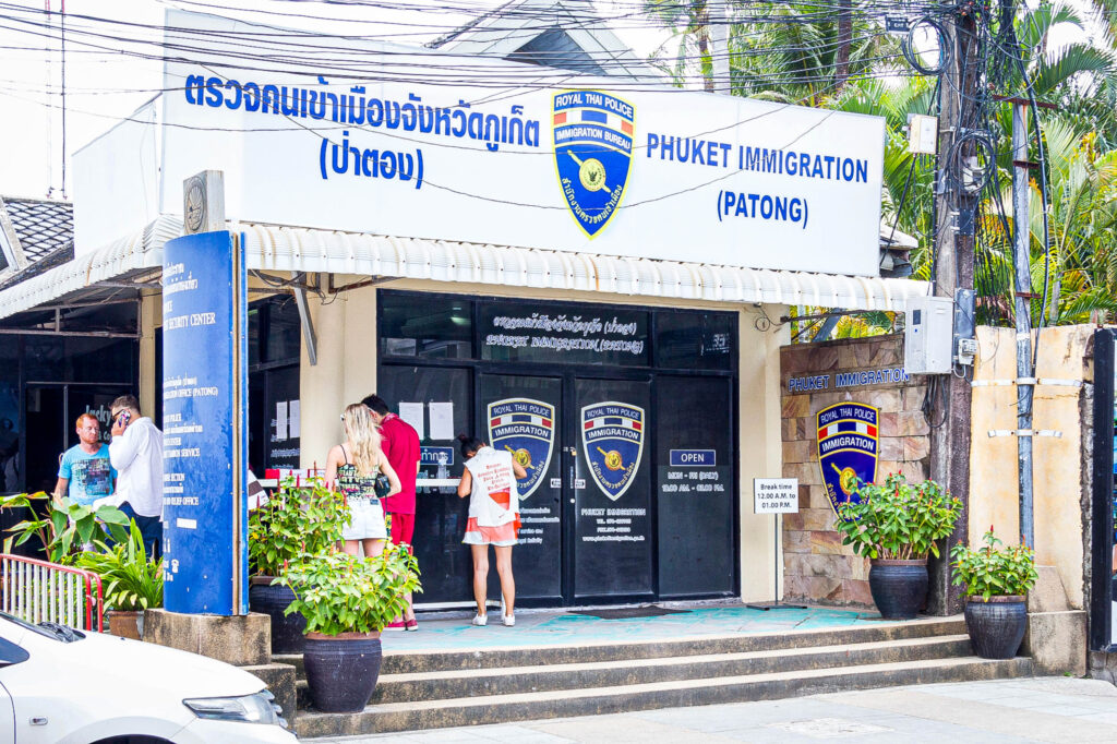 Außenansicht der Einwanderungsbehörde von Phuket (Patong) mit thailändischen und englischen Schildern. Mehrere Personen, die nach Thailand auswandern wollen, stehen vor dem Eingang, umgeben von Topfpflanzen und einem geparkten Fahrzeug.