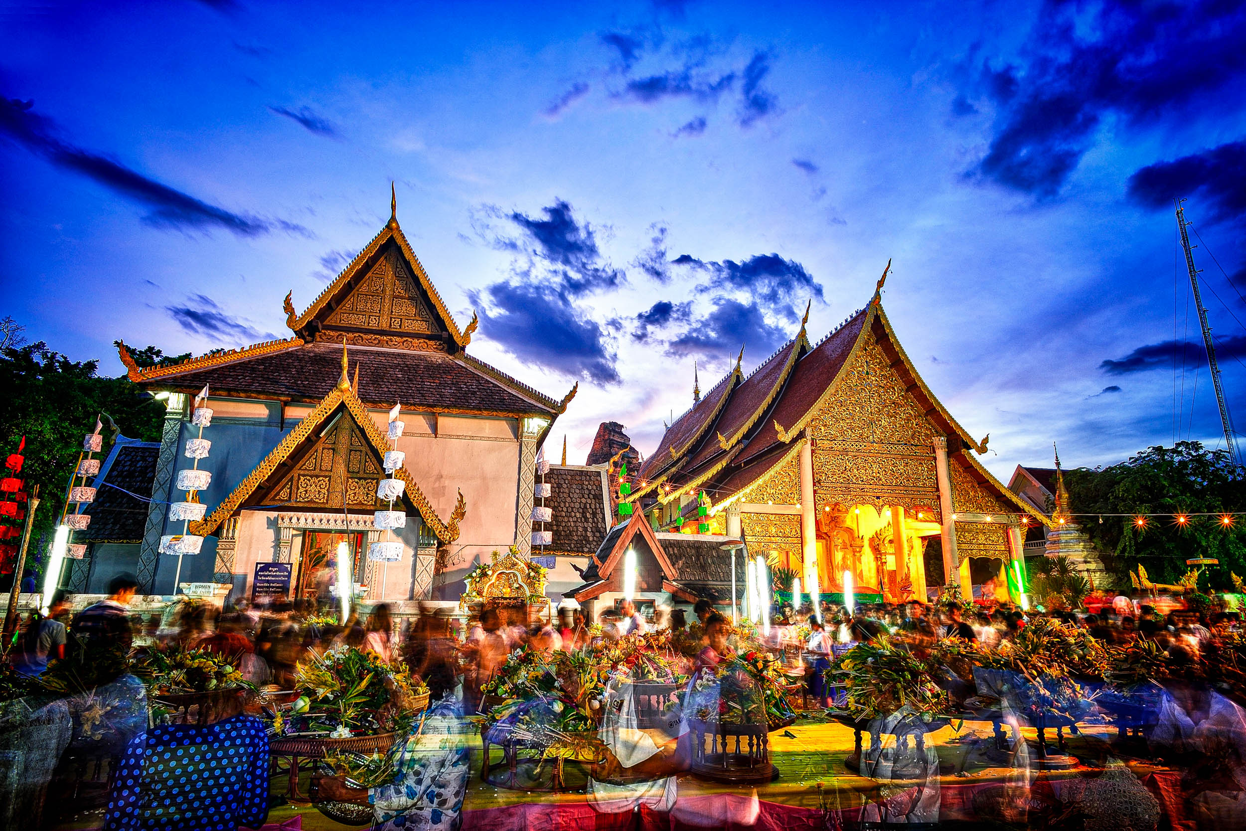 Eine lebhafte Festszene in einem traditionellen thailändischen Tempel in der Abenddämmerung, mit verzierten Dächern, bunten Lichtern und verschwommenen Menschenmengen, die sich um Tische versammeln und eine lebhafte und festliche Atmosphäre schaffen.