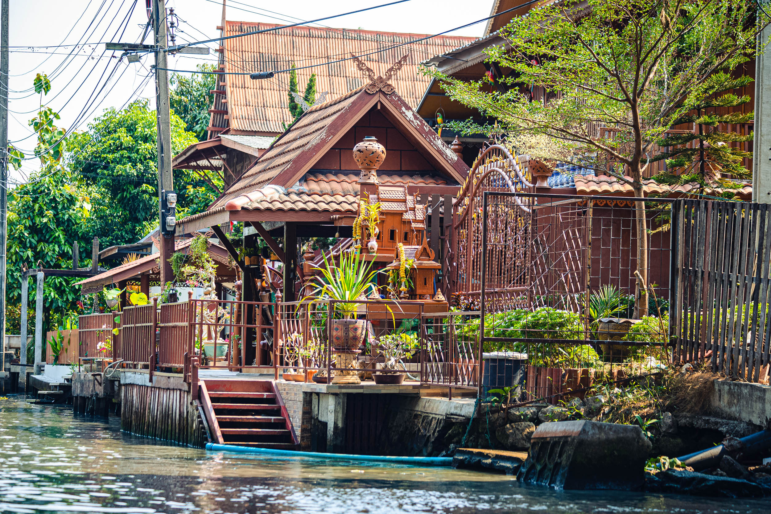 Traditionelle thailändische Holzhäuser mit verzierten Dächern und ein Geisterhaus liegen an einem Kanal, umgeben von üppigen Pflanzen und Bäumen, mit Stufen, die hinunter zum Wasser führen.