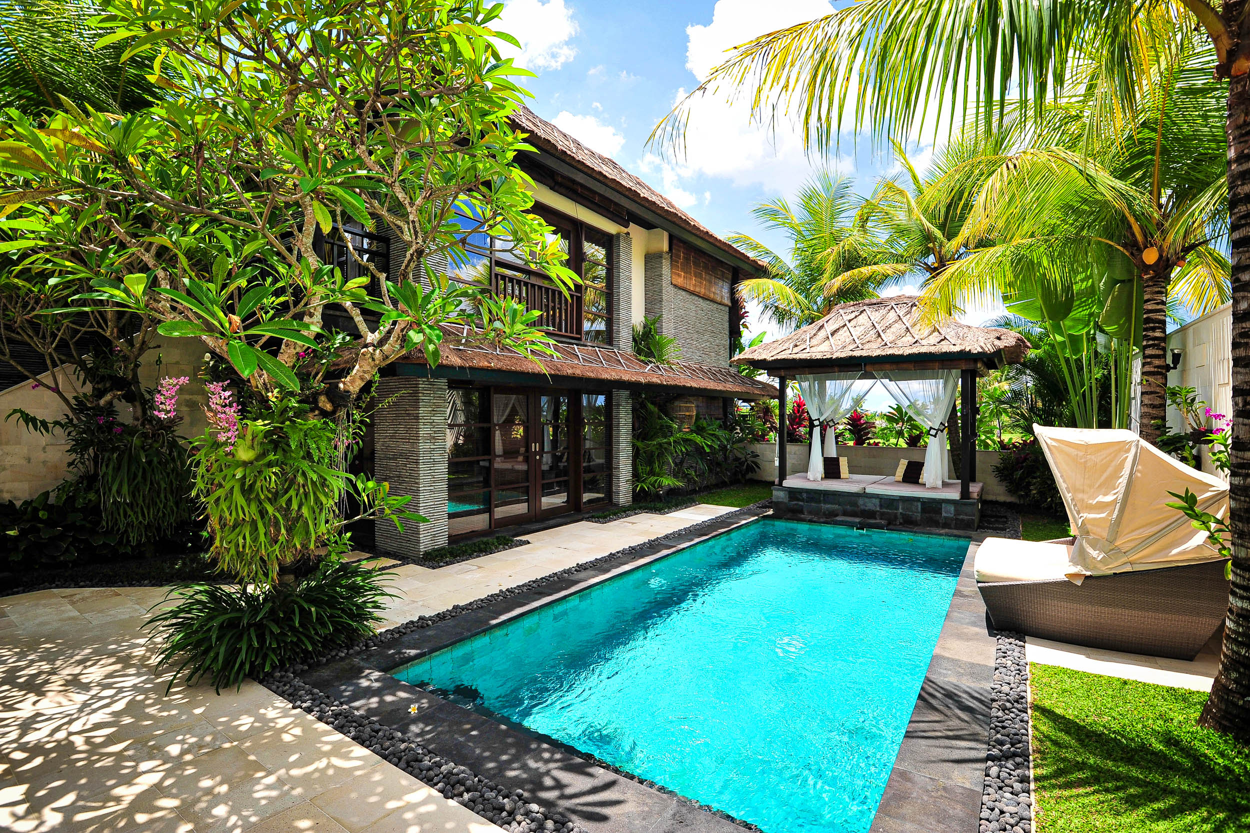 Eine tropische Villa mit einem privaten Pool, umgeben von üppigem Grün, Palmen und blühenden Pflanzen. Es gibt einen schattigen Lounge-Bereich neben dem Pool und große Glastüren, die zur Villa führen. Der Himmel ist hell und sonnig.