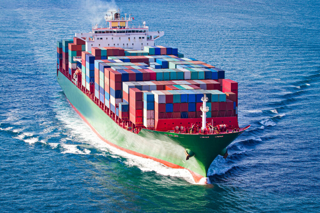 Ein großes Frachtschiff segelt über blaues Wasser in Richtung Thailand, voll beladen mit bunt gestapelten Schiffscontainern. Das Schiff erzeugt weiße Wellen, während es sich unter einem klaren Himmel vorwärts bewegt - ein inspirierender Anblick für alle, die vom Auswandern träumen.