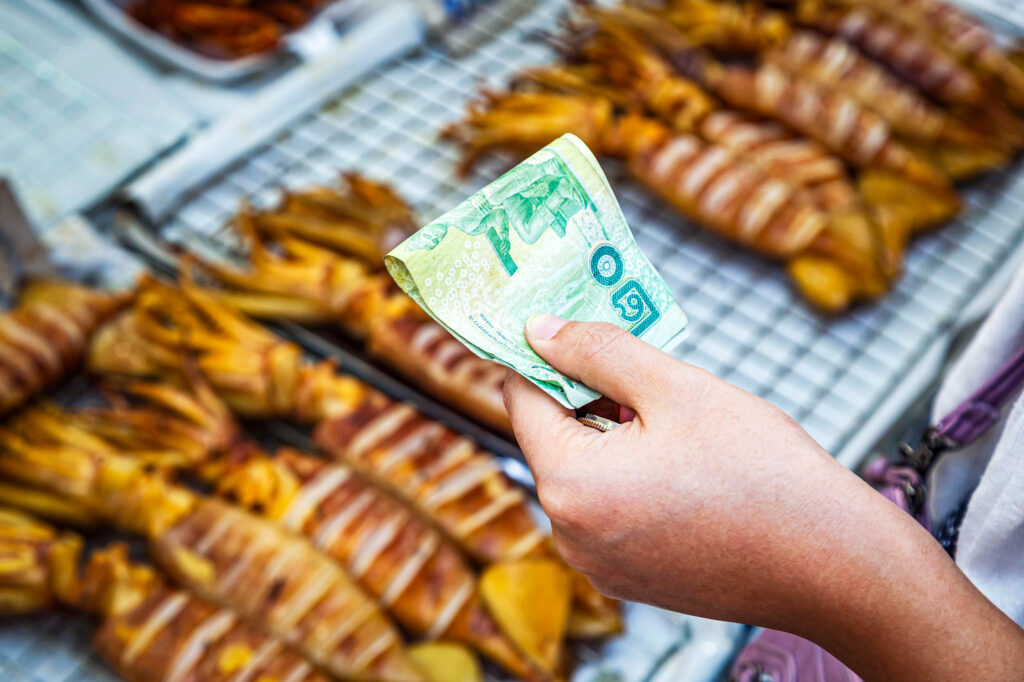 Eine Person hält thailändische Baht-Banknoten in der Hand und bereitet sich darauf vor, an einem Straßenstand zu bezahlen, an dem gegrillter Tintenfisch auf einem Metallgestell angeboten wird.