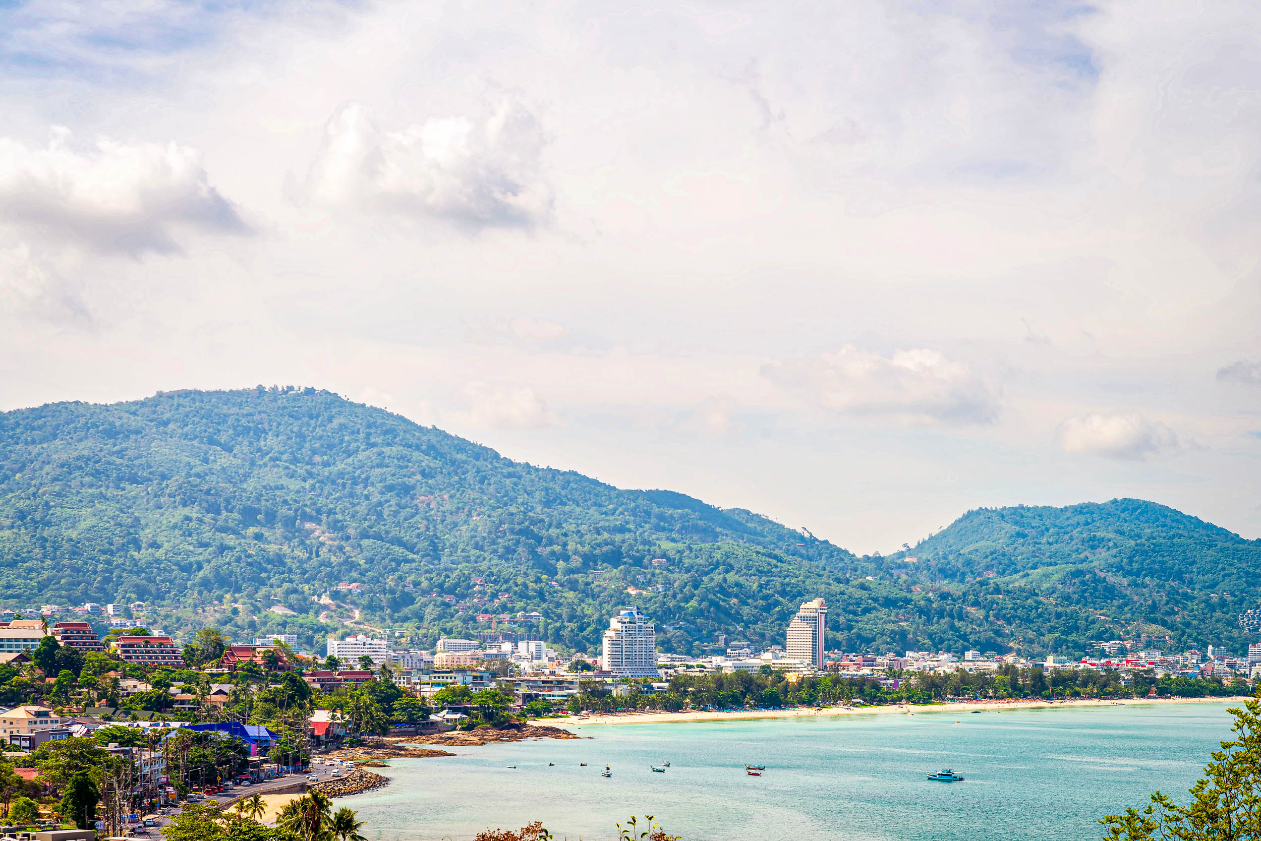 Eine Küstenstadt mit weißen Gebäuden und hohen Hotels liegt an einer türkisfarbenen Bucht, die von üppigen, grünen Bergen unter einem teilweise bewölkten Himmel umgeben ist. Kleine Boote treiben in der Nähe des Ufers.