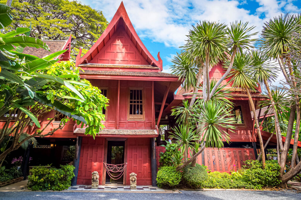 Ein traditionelles, rot gestrichenes Holzhaus im thailändischen Stil, umgeben von üppigen tropischen Pflanzen und Bäumen unter einem blauen Himmel mit Wolken - eine perfekte Kulisse für alle, die vom Auswandern nach Thailand träumen. Zwei Löwenstatuen bewachen den Eingang.