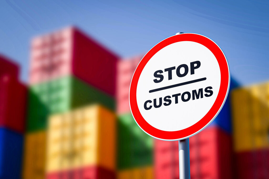 Ein STOP CUSTOMS-Schild im Vordergrund mit unscharfen bunten Schiffscontainern im Hintergrund, die vor einem blauen Himmel aufgestapelt sind und an die Reise derjenigen erinnern, die nach Thailand auswandern wollen.