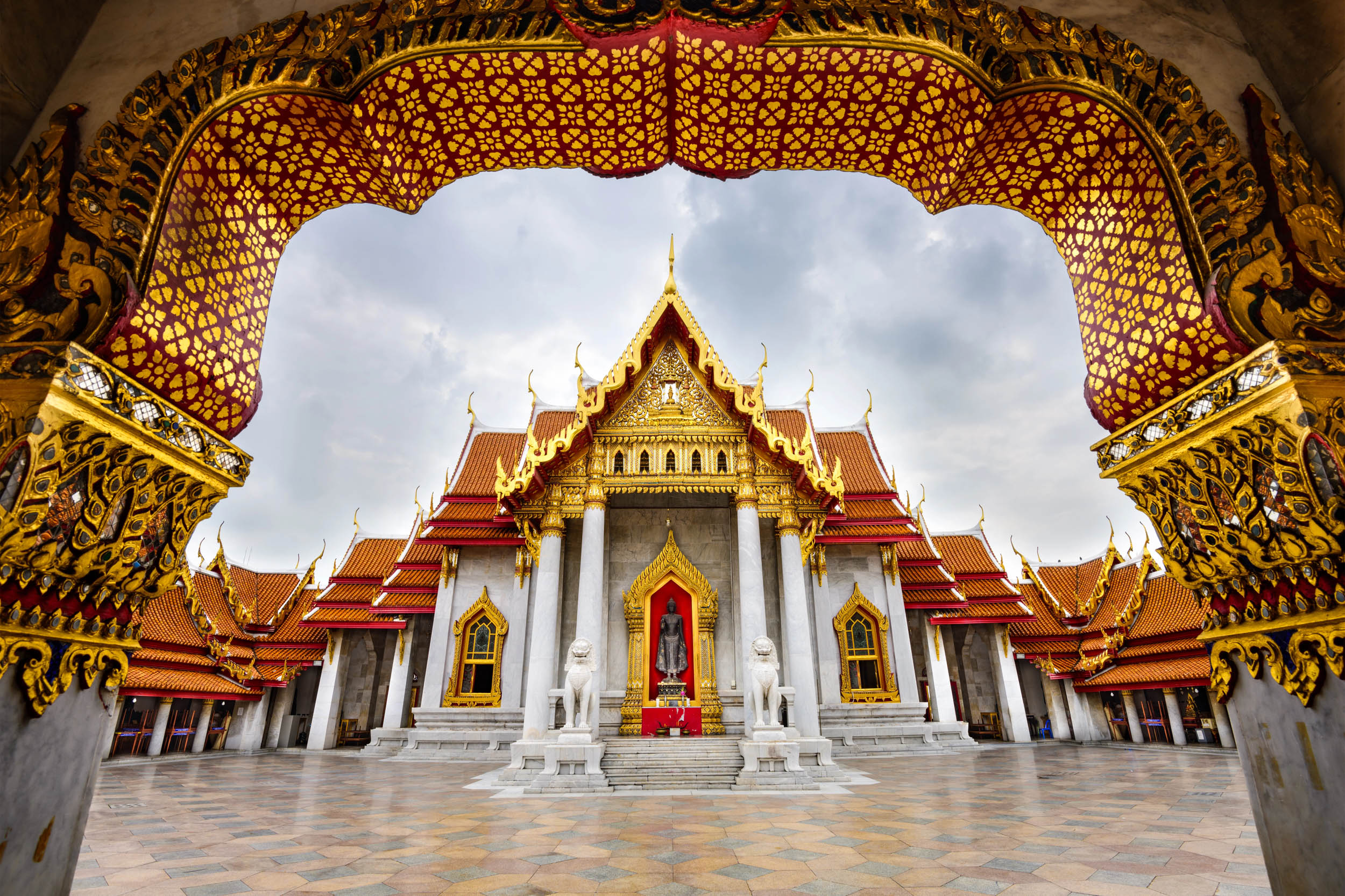 Buddhistischer Tempel mit verzierten goldenen Dachdetails, gewölbtem Eingang und zwei weißen Steinlöwen, die die Fassade bewachen. Die Szene wird von einem dekorativen Torbogen eingerahmt und steht unter einem bewölkten Himmel.
