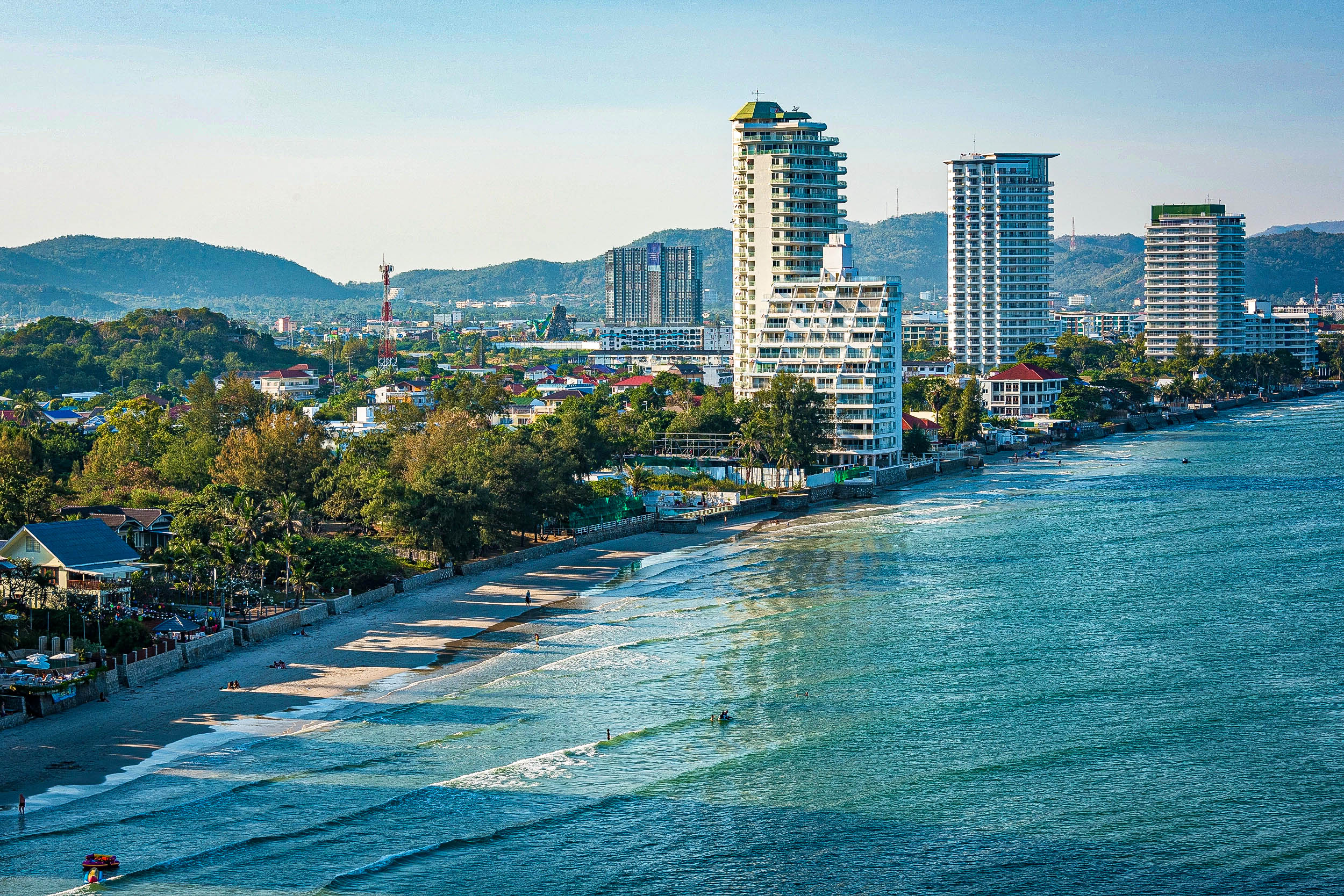 Eine Stadtlandschaft an der Küste mit hohen modernen Gebäuden, die einen Sandstrand säumen, sanften Wellen, die an die Küste klatschen, und grünen Hügeln im Hintergrund unter einem klaren blauen Himmel.