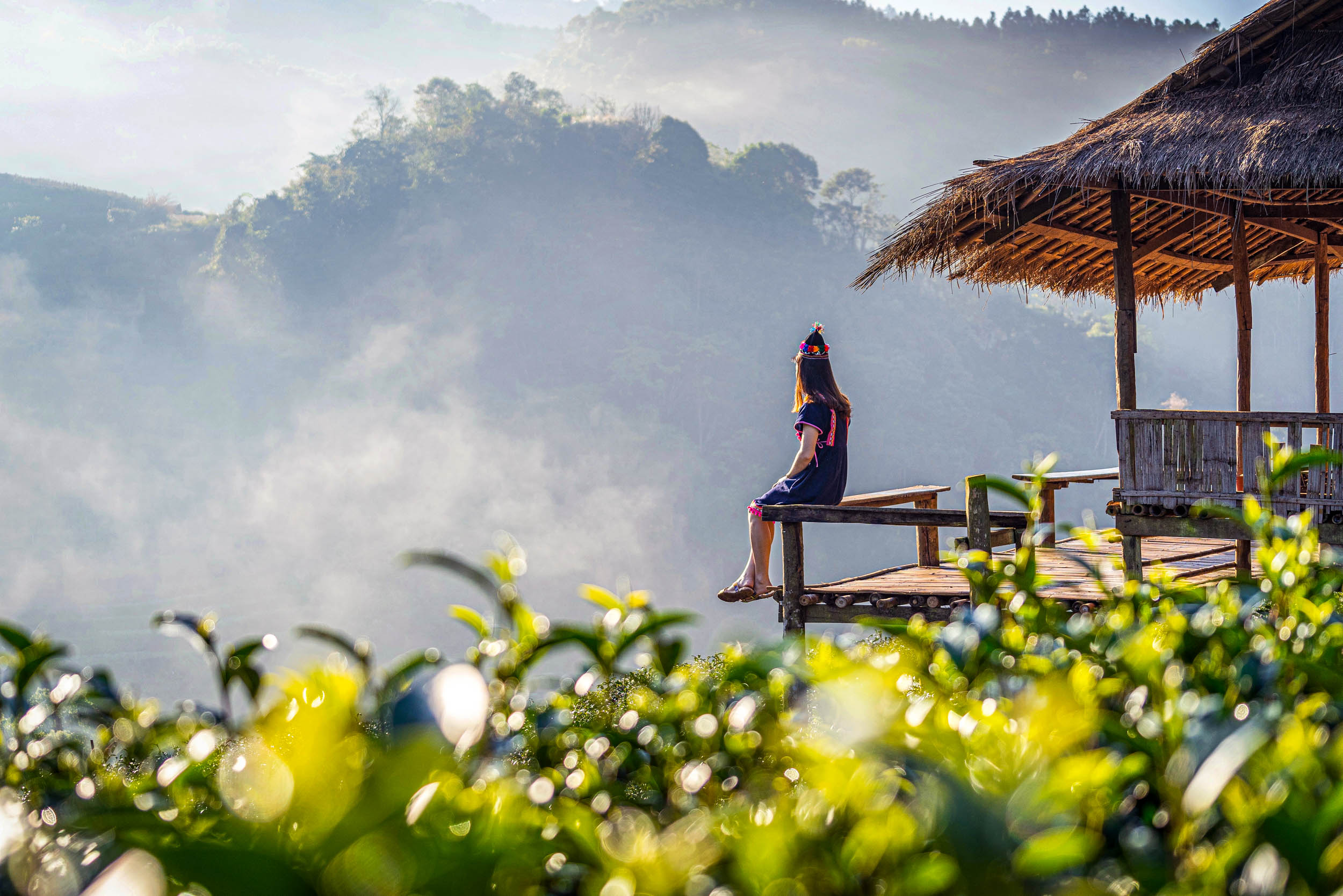 Eine Frau in traditioneller Kleidung sitzt am Rande einer Holzhütte mit Blick auf nebelgrüne Hügel und Teepflanzen und genießt die Aussicht auf die Berge im sanften Morgenlicht.